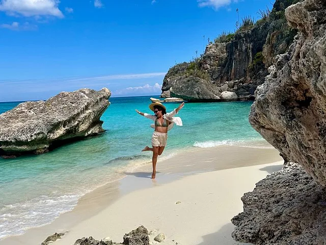 Una mujer saltando de alegría en una playa de arena blanca cerca de grandes rocas.